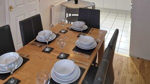 A dining area with a wooden table set for four, white plates, and wine glasses. There are black chairs, a small potted plant, and a window with a view of a brick building. - HEREFORD – Finch House: Modern 4-Bed, Dog Friendly, Sleeps 10 + Pool Table (Hereford)