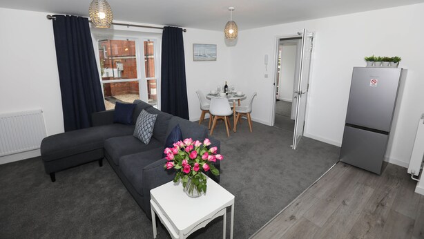 Un salon moderne avec un canapé gris, une table blanche ornée d’un vase de fleurs roses, un coin repas avec une table et des chaises blanches, et un réfrigérateur gris.