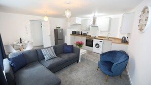 Un salon moderne avec un canapé d’angle gris, un fauteuil bleu et une petite table blanche sur laquelle est posé un vase de fleurs. On aperçoit, au fond, une cuisine avec des meubles blancs, un four à micro-ondes et un lave-linge.