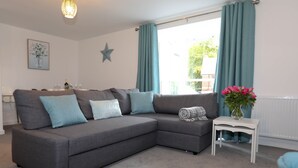 A modern living room with a grey sectional sofa, a small white side table, and a window with teal curtains. - EXMOUTH, DEVON - Apt 2, Strand House: 2 Bed, Central Location, Sleeps 6, (Exmouth)
