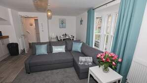 A modern living room with a grey sectional sofa, a small white table with a vase of pink flowers, and a dining area in the background. - EXMOUTH, DEVON - Apt 2, Strand House: 2 Bed, Central Location, Sleeps 6, (Exmouth)