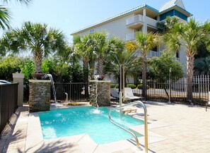 A swimming pool with a stone wall and a metal railing, surrounded by palm trees and a fence.