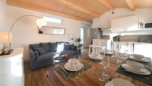 Un salon moderne avec un plafond en bois, un canapé gris, une table à manger dressée avec de l’assiette et des verres blancs, et un coin cuisine doté de meubles blancs et d’un four à micro-ondes.