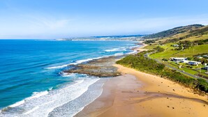 A coastal landscape with a sandy beach, ocean waves, and a road curving along the coastline. - Frankie - A wonderful place to escape the city (Skenes Creek)