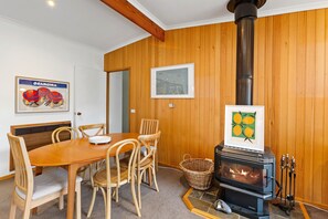 A wooden-paneled room with a dining table, chairs, a framed picture, and a wood-burning stove. - Frankie - A wonderful place to escape the city (Skenes Creek)