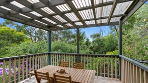 A wooden deck with a table and chairs, surrounded by a metal railing and lush greenery.