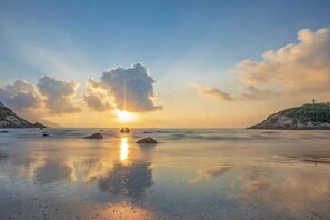 A beach scene with the sun setting over the water, rocks in the foreground, and hills in the background. - Jingangwan Hotel (Taishan)