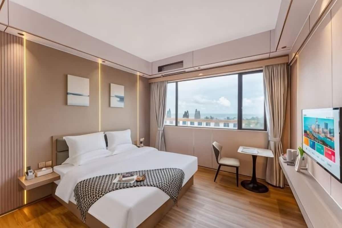 A modern hotel room with a large bed, bedside tables, a small desk, a chair, a TV, and a window with a city view.