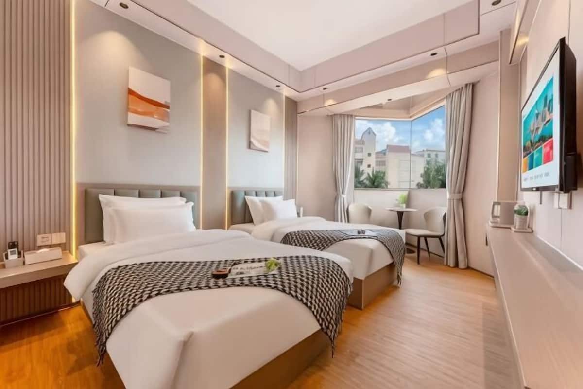 A hotel room with two beds, a small table, a TV, and a window with a view of buildings.