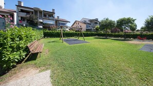 A well-maintained grassy area with a wooden swing set and a bench, surrounded by neatly trimmed hedges and a row of residential buildings in the background. - Lauri 0/18 by Wonderful Italy (Desenzano del Garda)