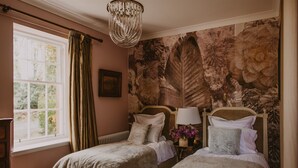 A bedroom with two beds, a window with curtains, a chandelier, and a floral wallpaper.