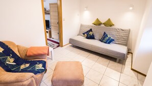 Una sala de estar con un sofá, una silla y un pequeño reposapiés. Se ve una puerta que conduce a otra habitación.