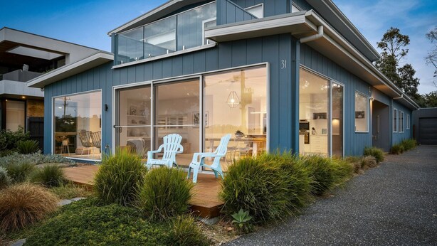 Une maison moderne dotée de grandes baies vitrées, d’une terrasse en bois et d’un espace salon extérieur.