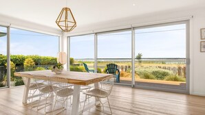 Une pièce spacieuse avec une grande table en bois et des chaises en treillis métallique, de grandes portes-fenêtres coulissantes donnant sur une vue sur une plage et une végétation luxuriante.