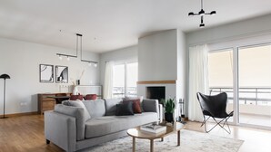 A modern living room with a grey sofa, a coffee table, and a black chair. There is a fireplace and a large window with curtains. - Celestial 3BR Apartment with Balcony (Palaio Faliro)