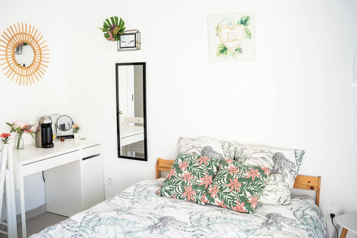 A bedroom with a bed, a desk, a mirror, and a wall-mounted plant.