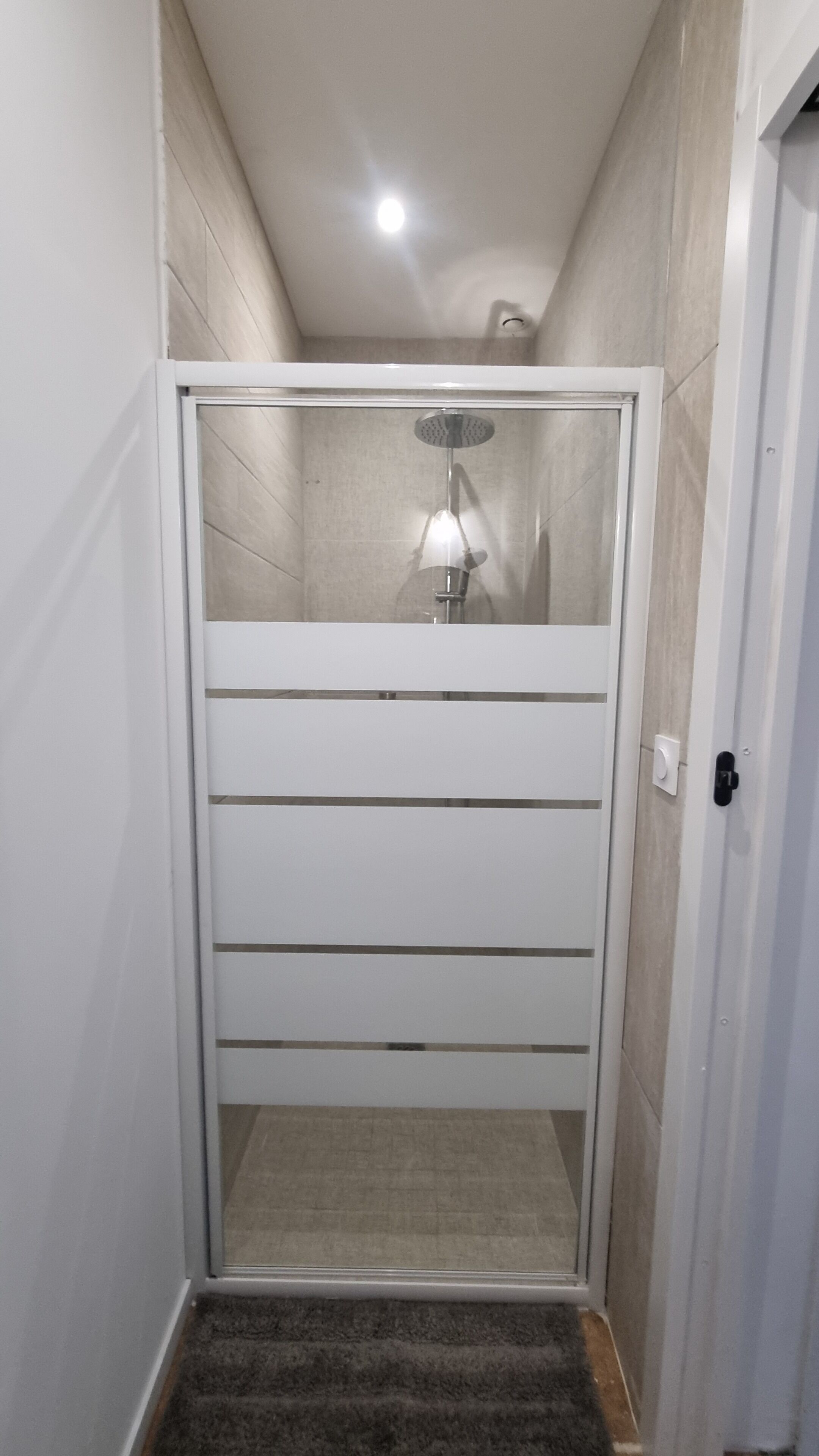 A narrow hallway leading to a shower area with a glass door, a light fixture above, and a door on the right.