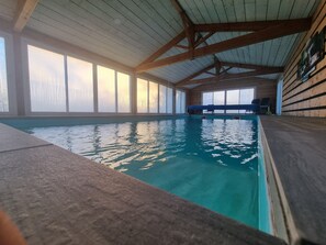 Una piscina interior con agua cristalina, vigas de madera en el techo y grandes ventanales que dejan entrar luz natural.