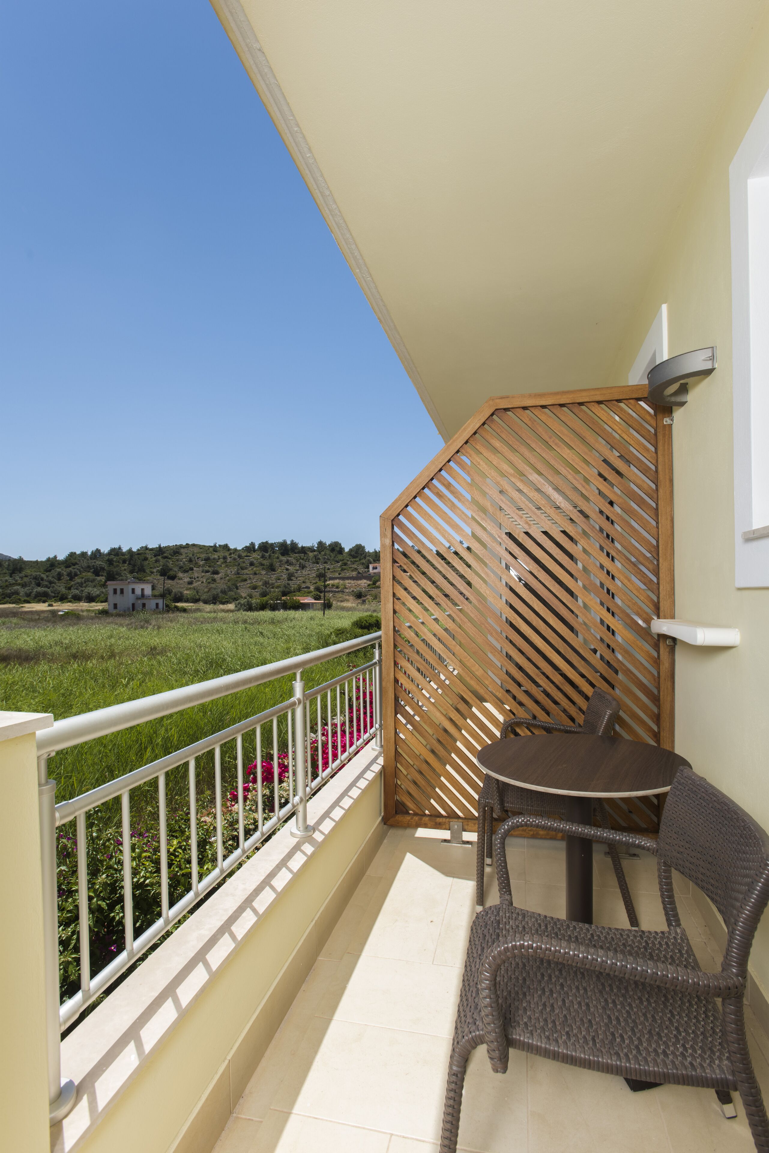 Un balcón con una mesa, una silla, un panel de madera y vistas a un edificio y campo a lo lejos.