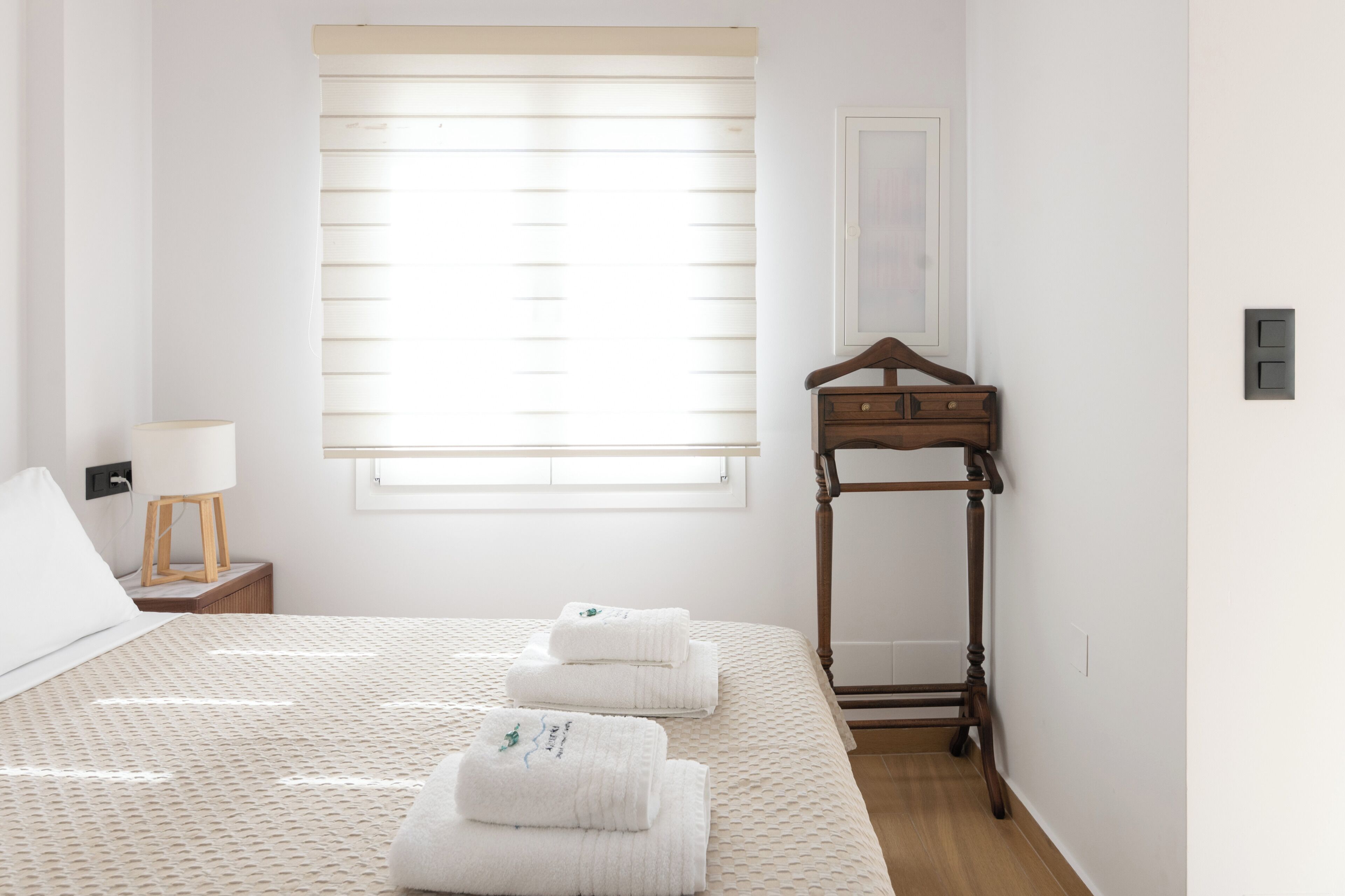 A hotel room with a bed, a bedside table with a lamp, a wooden bedside table, and a window with blinds.