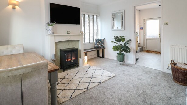 A living room with a fireplace, a television mounted above it, a dining table with chairs, and a large window with blinds.