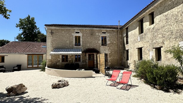 A building with a stone facade, a tiled roof, and a gravel courtyard with a red chair and a barbecue grill. - Charming gîte "Chez Erwann" 108m2 (Saint-Just)