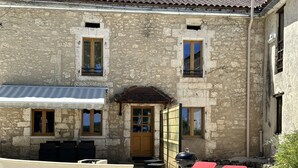 A stone building with a wooden door, two windows, and a covered patio area with two red chairs. - Charming gîte "Chez Erwann" 108m2 (Saint-Just)
