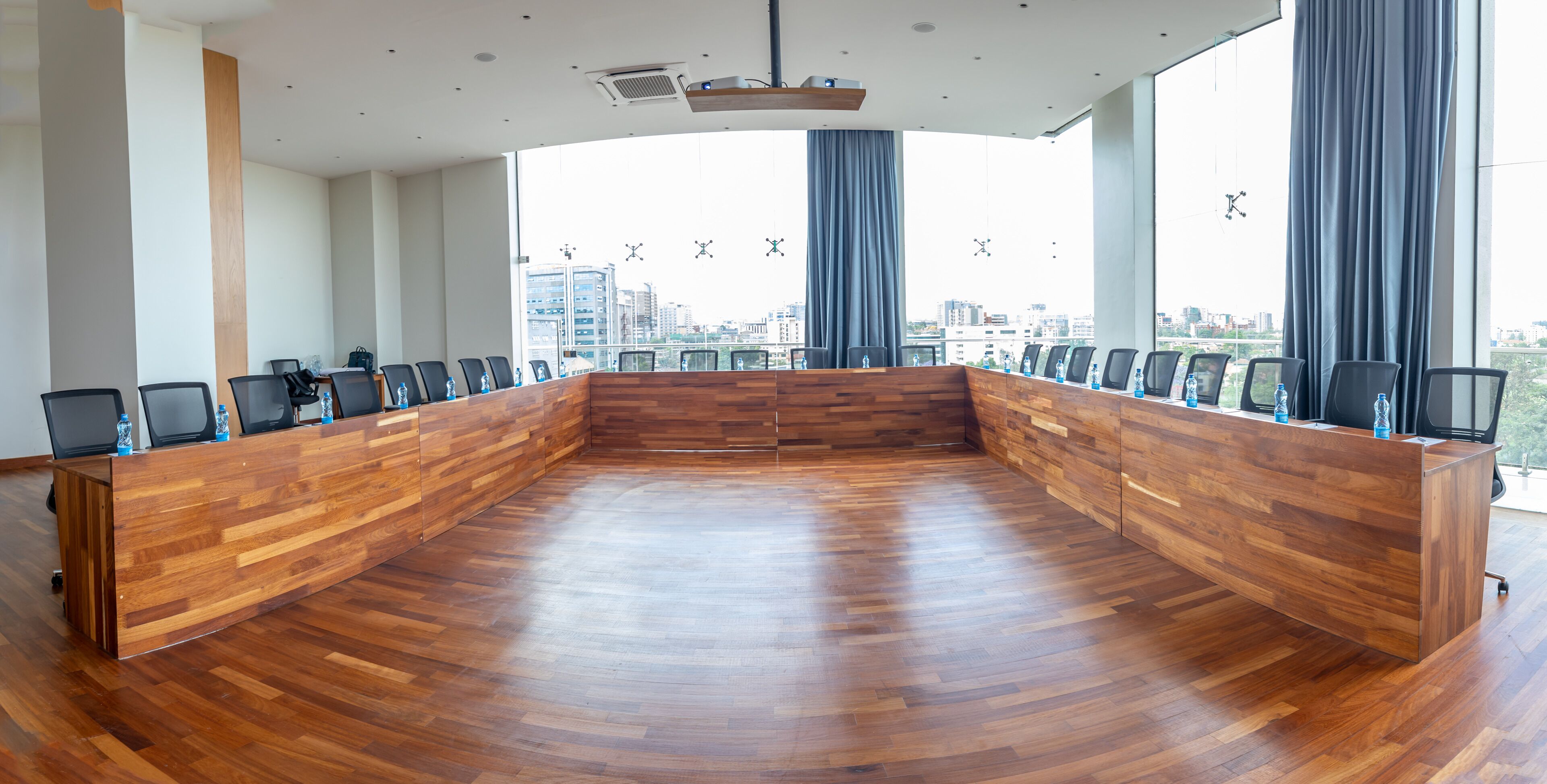 Een vergaderruimte met een houten tafel en stoelen, grote ramen voorzien van gordijnen en een uitzicht op de stad.