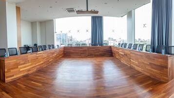 Een vergaderruimte met een houten tafel en stoelen, grote ramen voorzien van gordijnen en een uitzicht op de stad.
