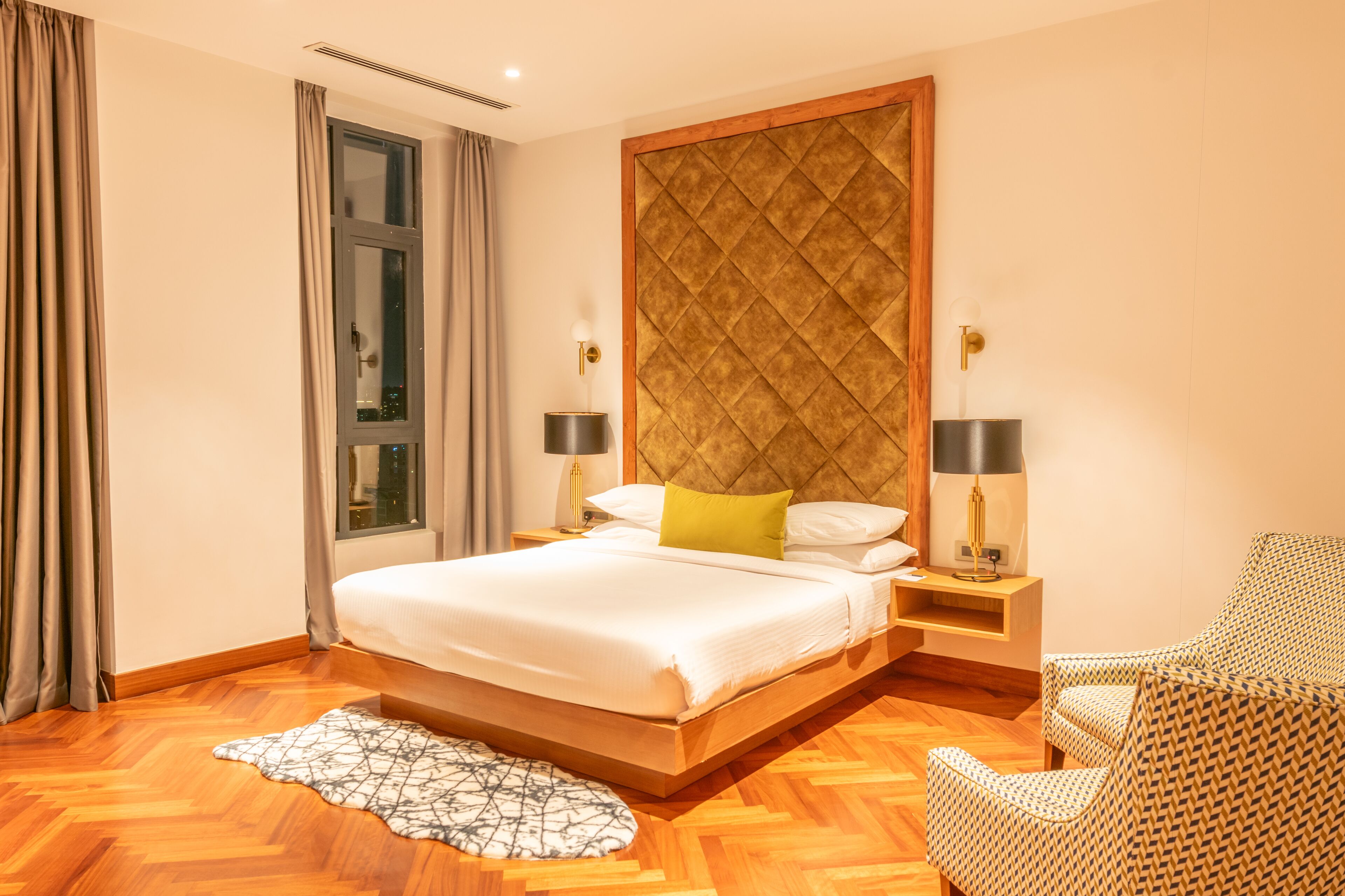 Quarto de hotel com uma cama grande, duas mesas de cabeceira com abajures, uma cadeira estampada e uma janela com cortinas.