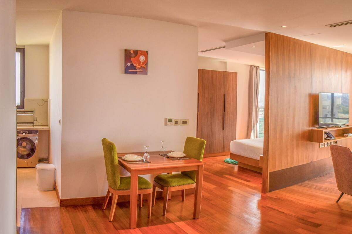 A modern hotel room with a wooden floor, a dining area with green chairs, a bed, a TV, and a view of the city.