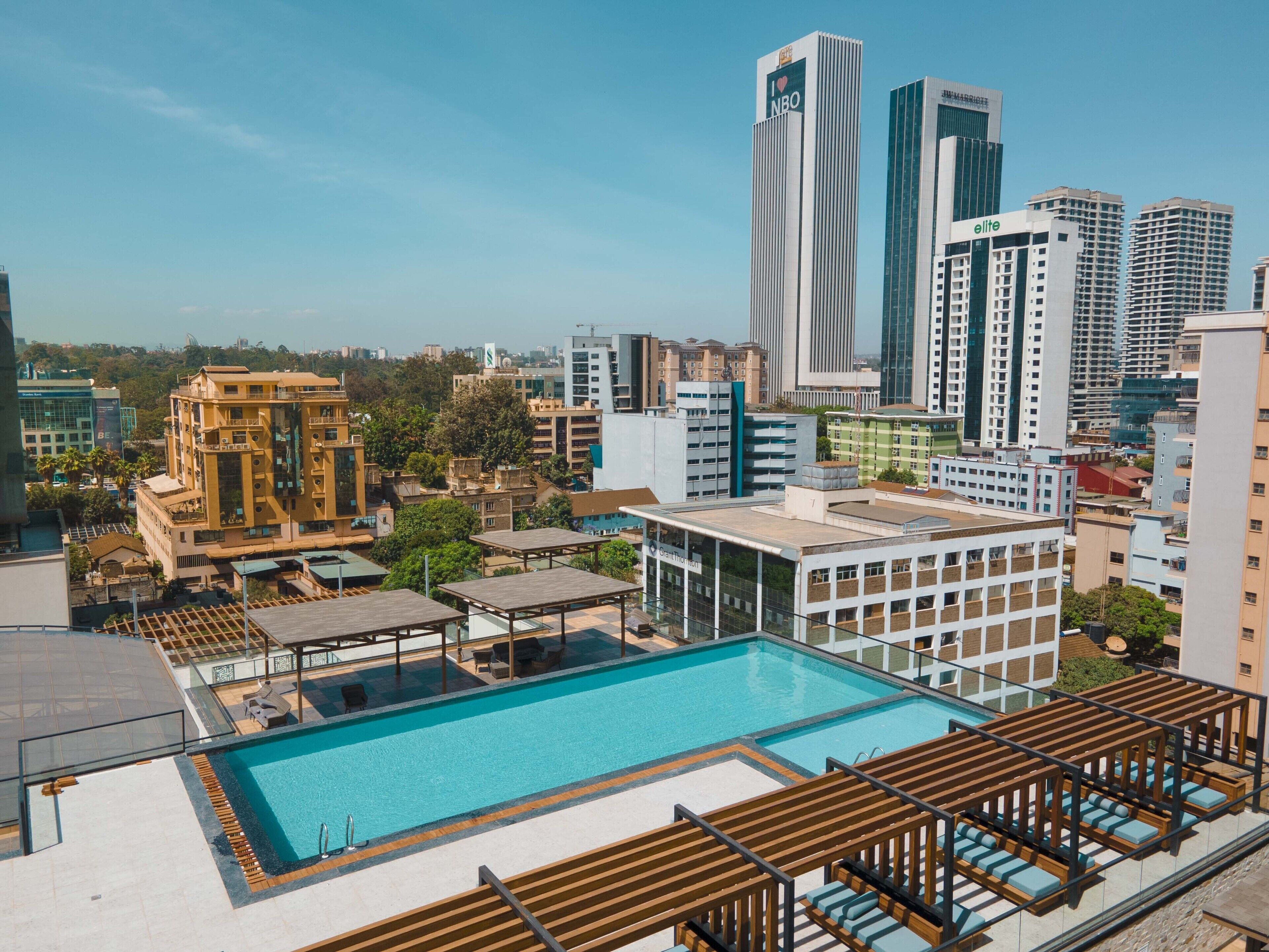 A rooftop pool with wooden pergolas and lounge chairs, surrounded by modern buildings and a clear sky.
