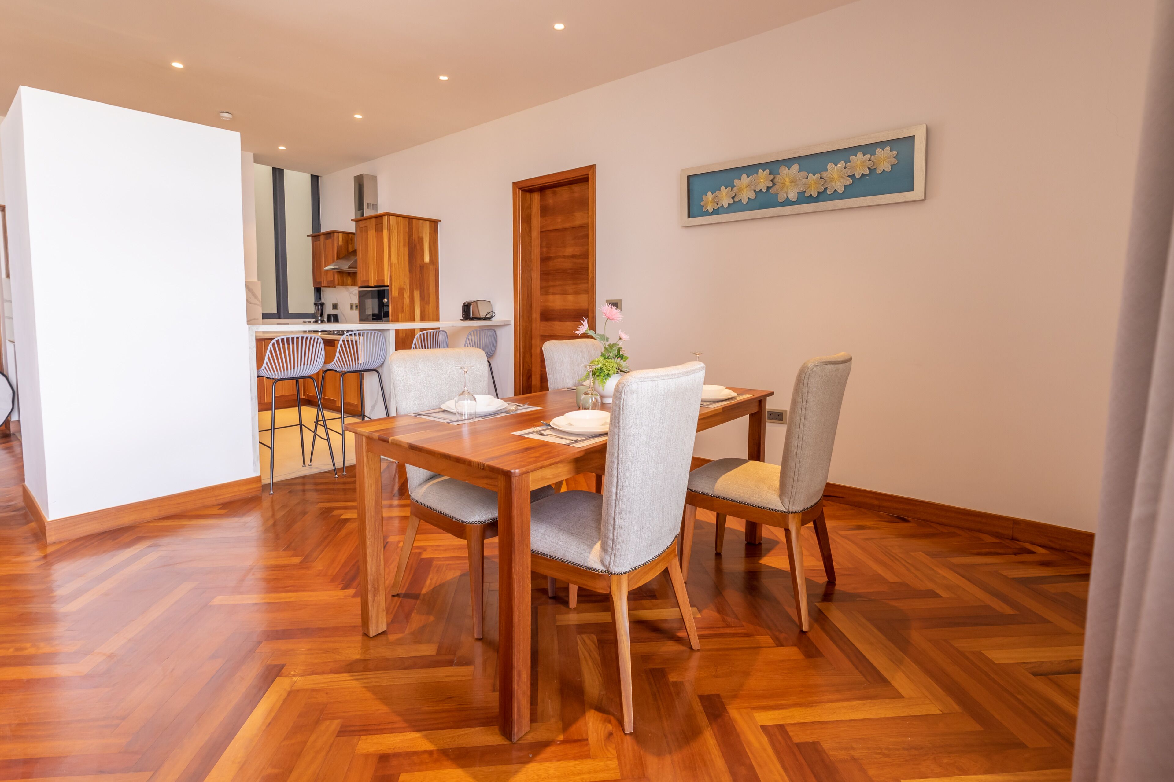 A dining area with a wooden table and chairs, a kitchen in the background, and a wall with a floral painting.