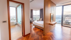 A modern hotel room with a large bed, a seating area with two chairs, a view of the city through large windows, and a wooden floor.