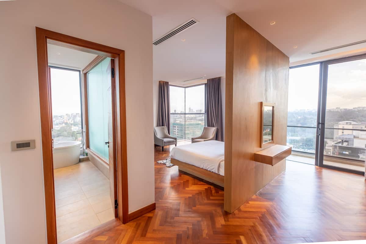 A modern hotel room with a large bed, a seating area with two chairs, a view of the city through large windows, and a wooden floor.