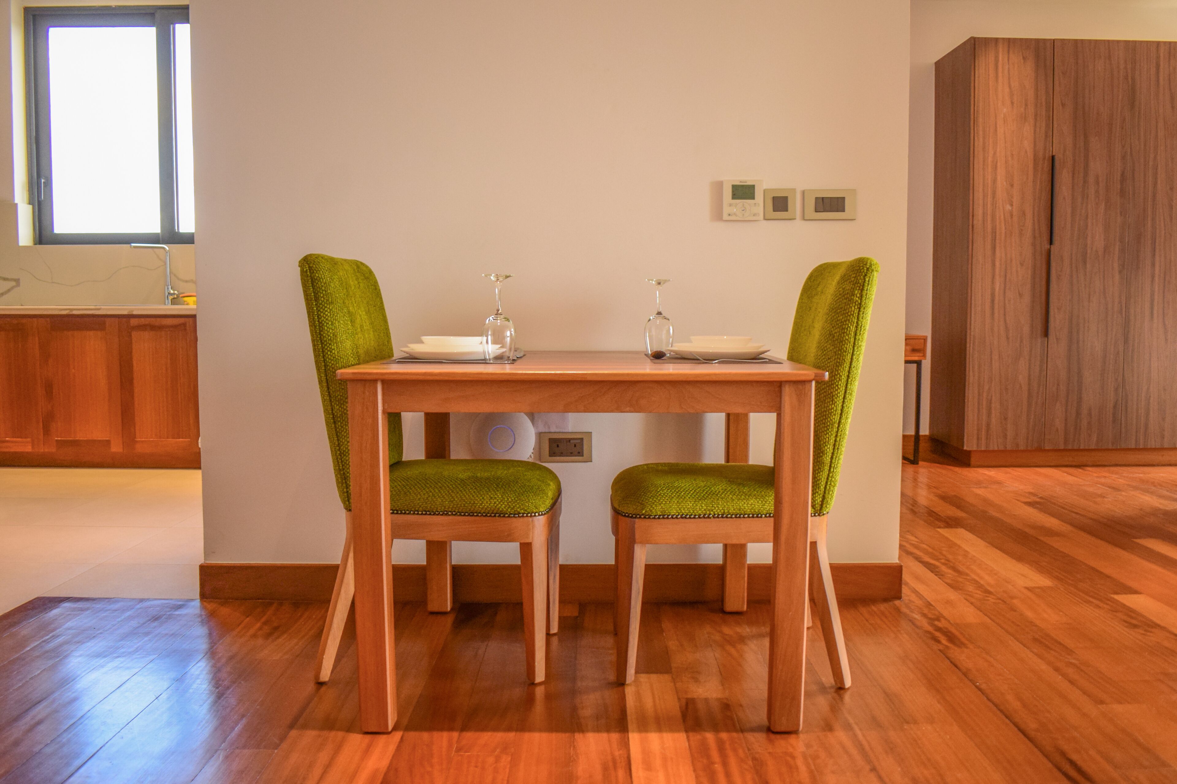 Un'area pranzo con un tavolo di legno e due sedie rivestite in verde, una finestra con persiane e un armadietto di legno.