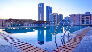 A swimming pool with a wooden deck and metal railings, surrounded by high-rise buildings and a clear sky.
