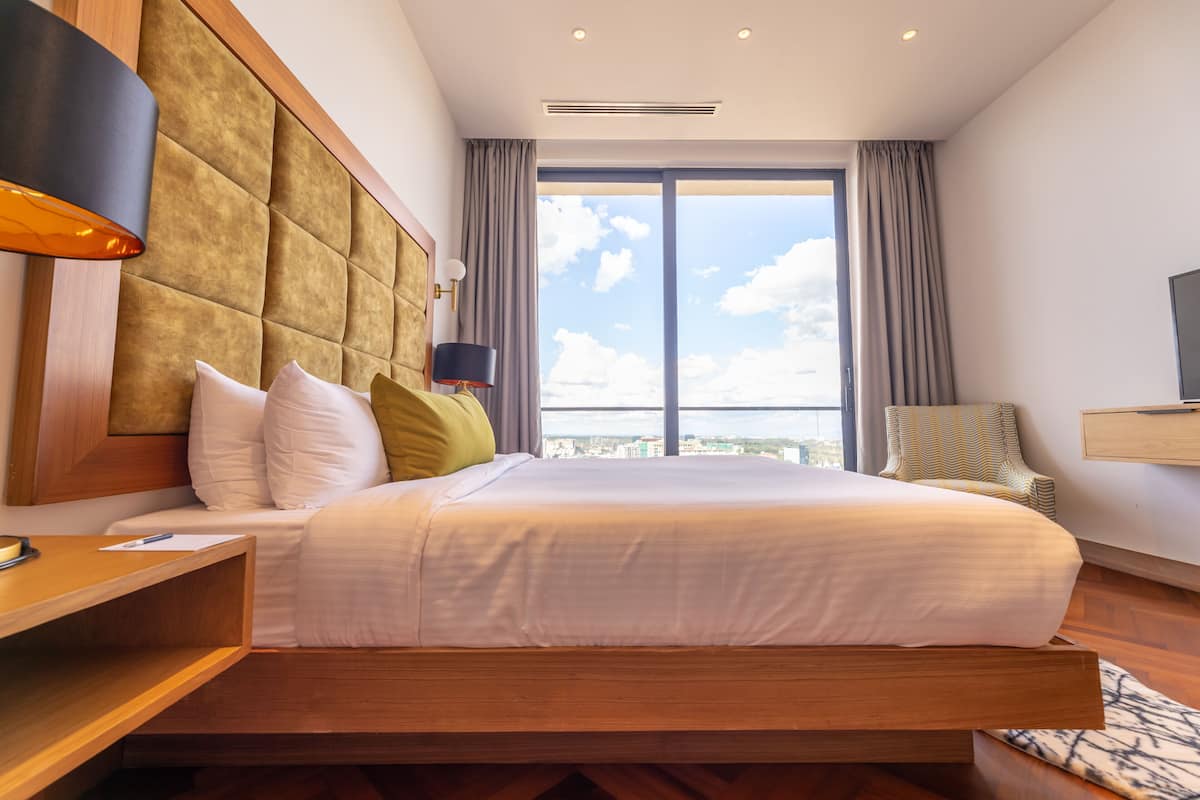 A modern hotel room with a large bed, a wooden headboard, a desk, a chair, and a large window with a city view.