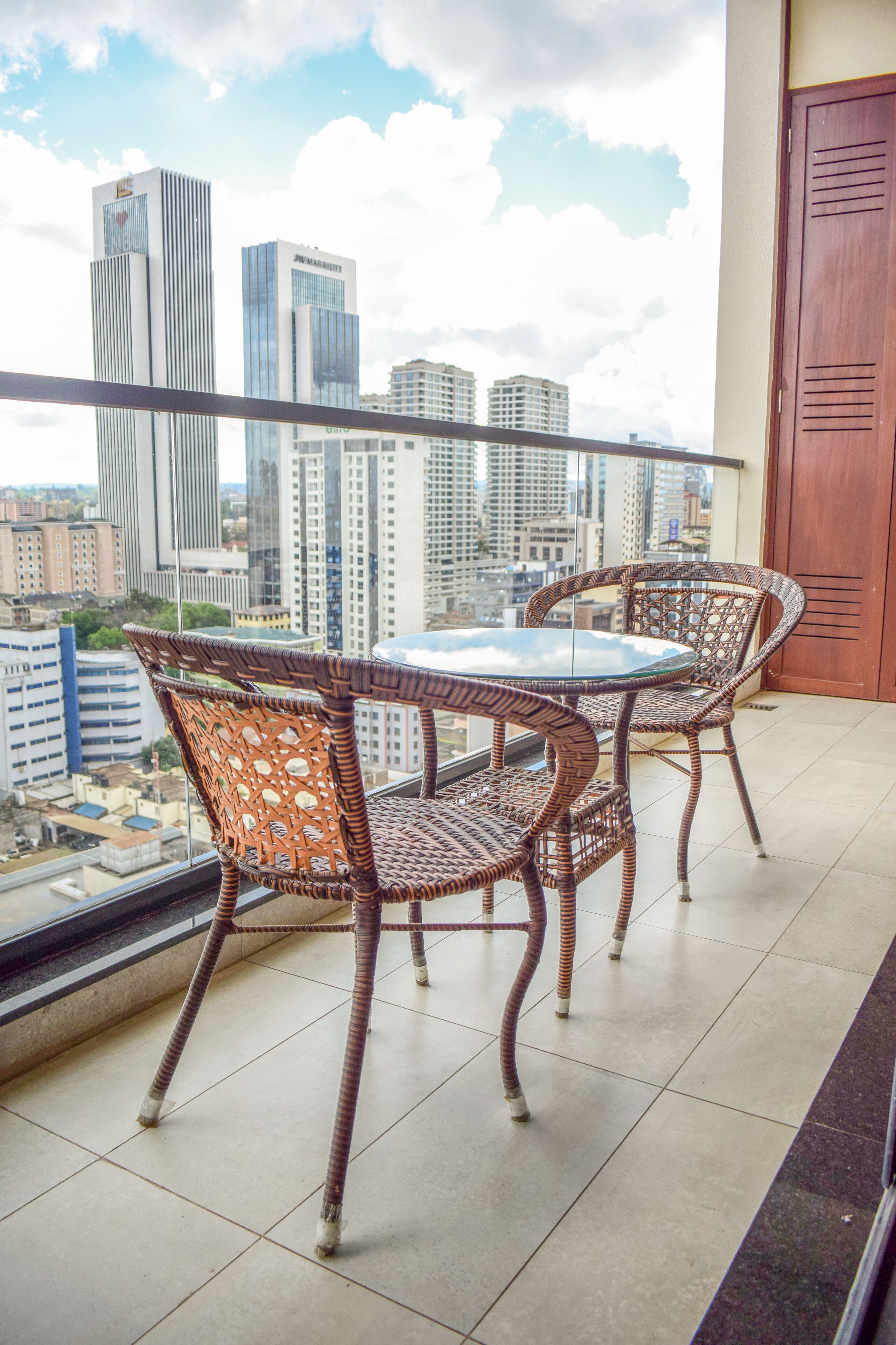 A balcony with wicker chairs and a glass table, overlooking a cityscape with tall buildings.