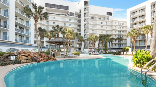 Una piscina rodeada de sillas de descanso y palmeras, con un edificio de varios pisos al fondo.