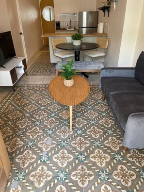 Un salon avec un sol à motifs, une table basse ronde en bois agrémentée d’une plante, un canapé gris et un coin repas avec une table et des chaises.