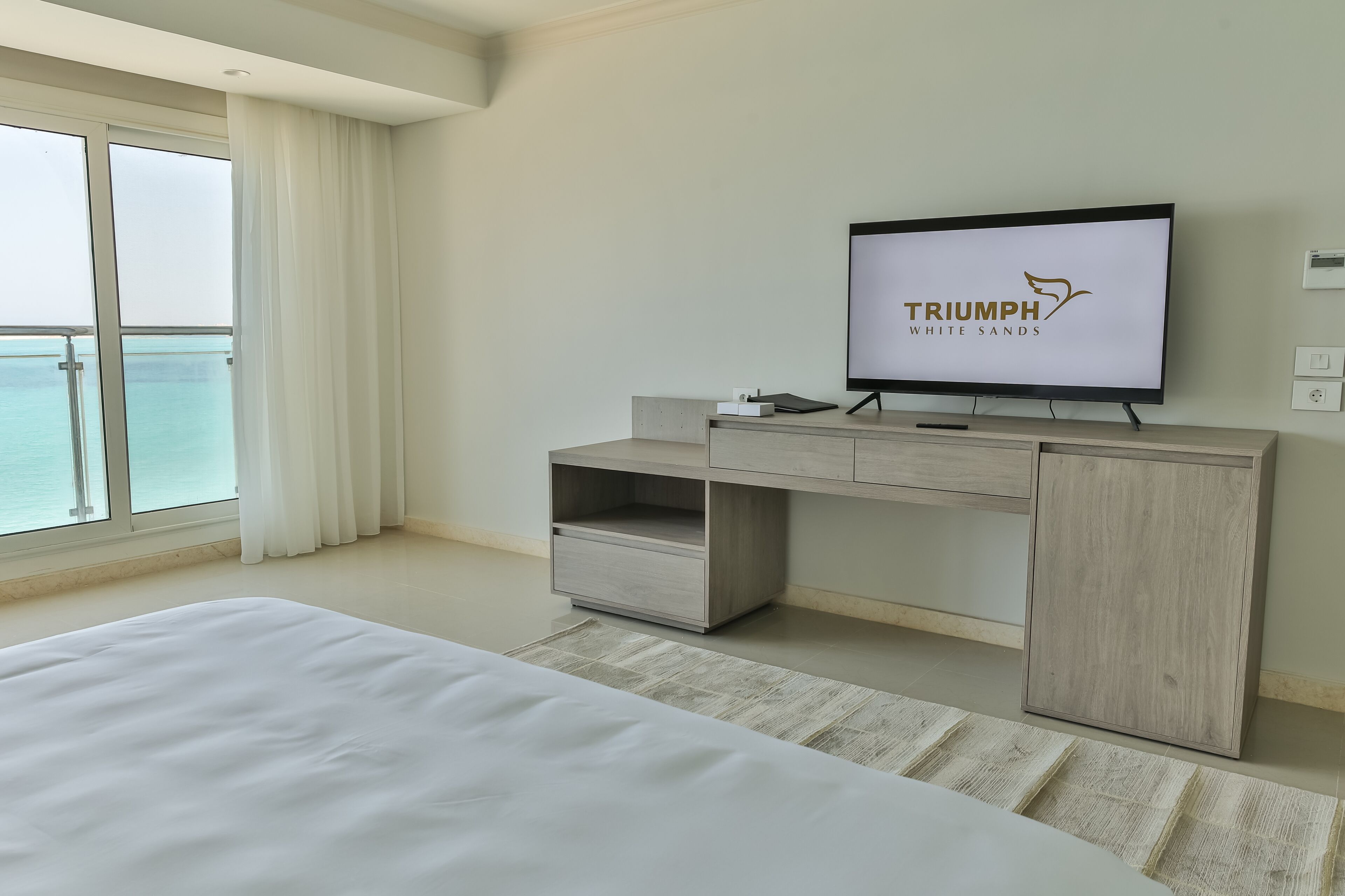 Una habitación de hotel moderna con una cama grande, un televisor mostrando 'TRIUMPH WHITE SANDS', un escritorio de madera y vistas al mar a través de amplios ventanales.