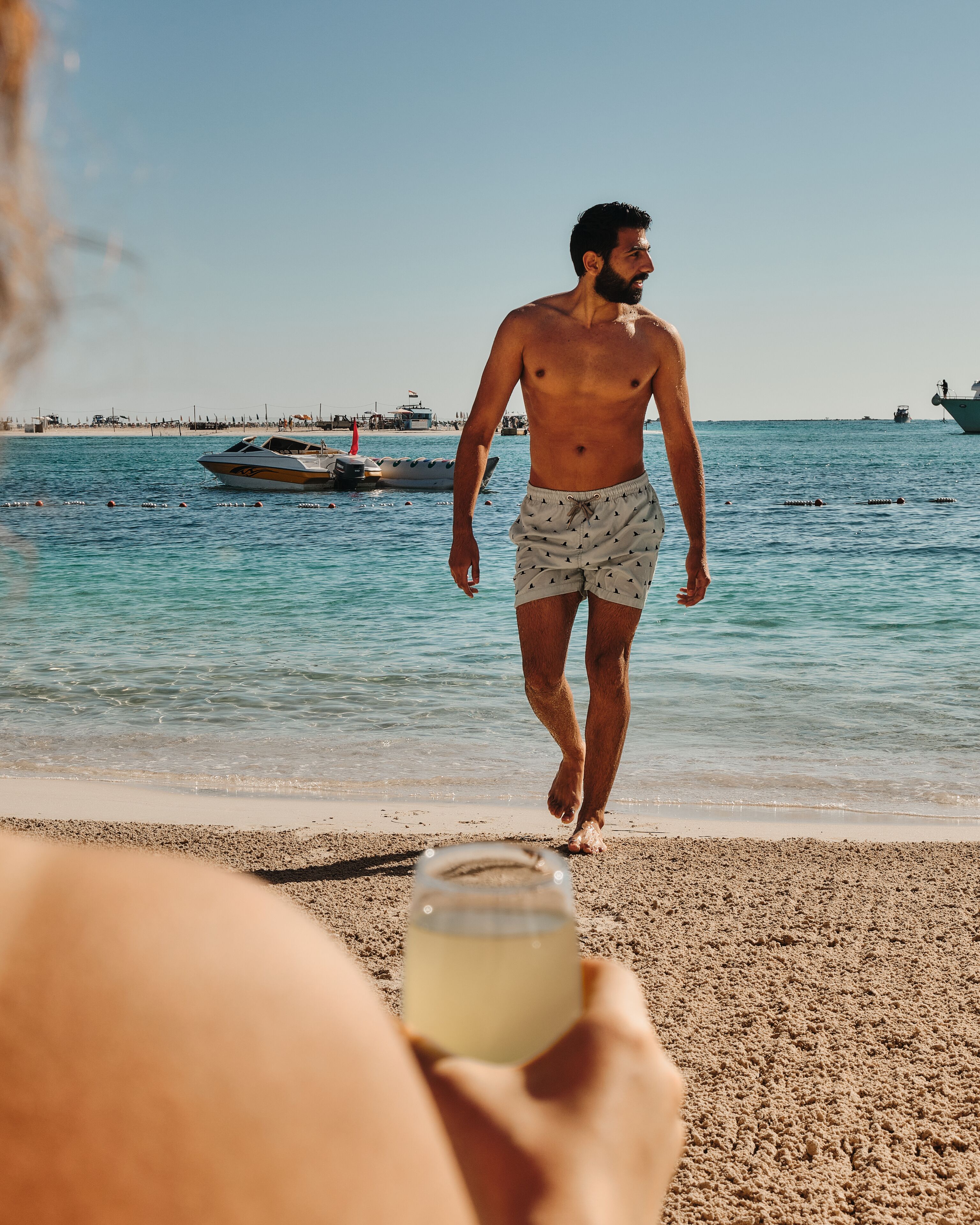 Une scène de plage avec une personne qui marche en direction de l’eau, des bateaux en arrière-plan et un ciel dégagé.