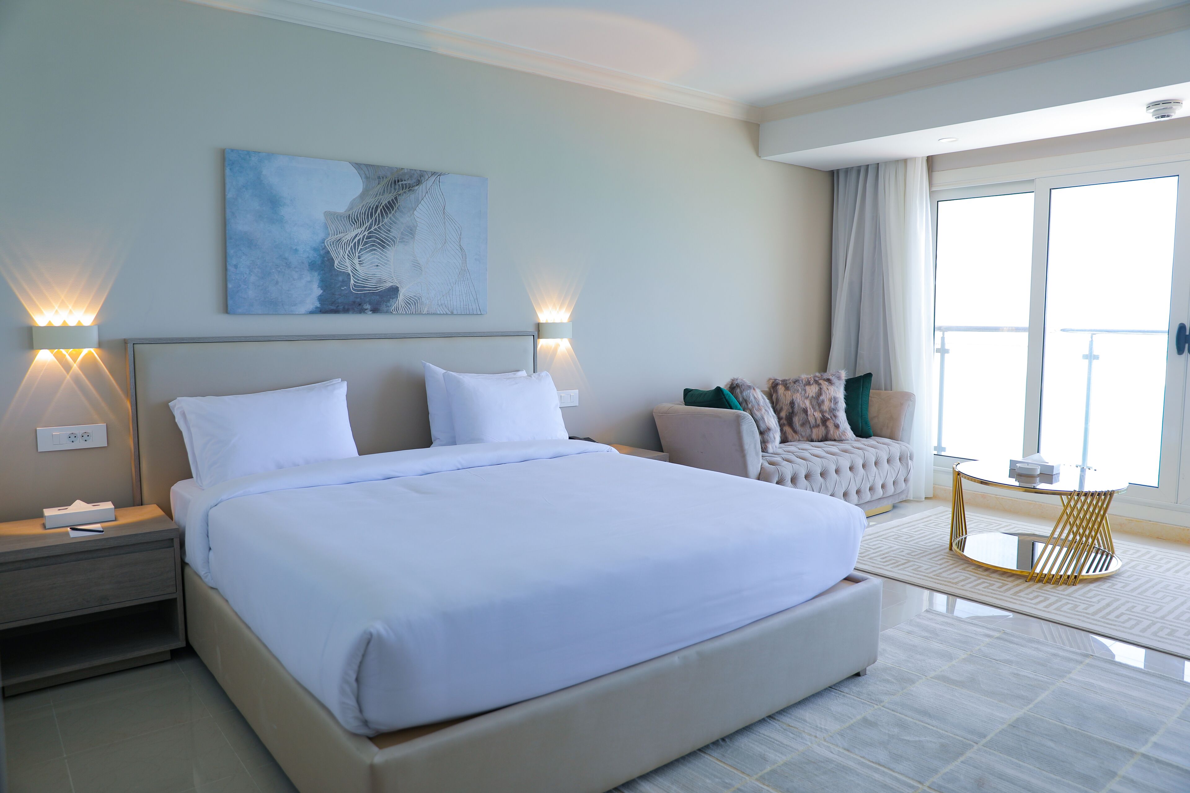 A modern hotel room with a large bed, bedside lamps, a sofa, a small round table, and a large window with curtains.