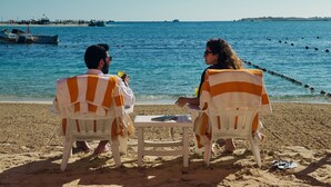 Zwei Personen sitzen an einem Tisch an einem sandigen Strand, im Hintergrund ein klarer, blauer Himmel und ruhige See.