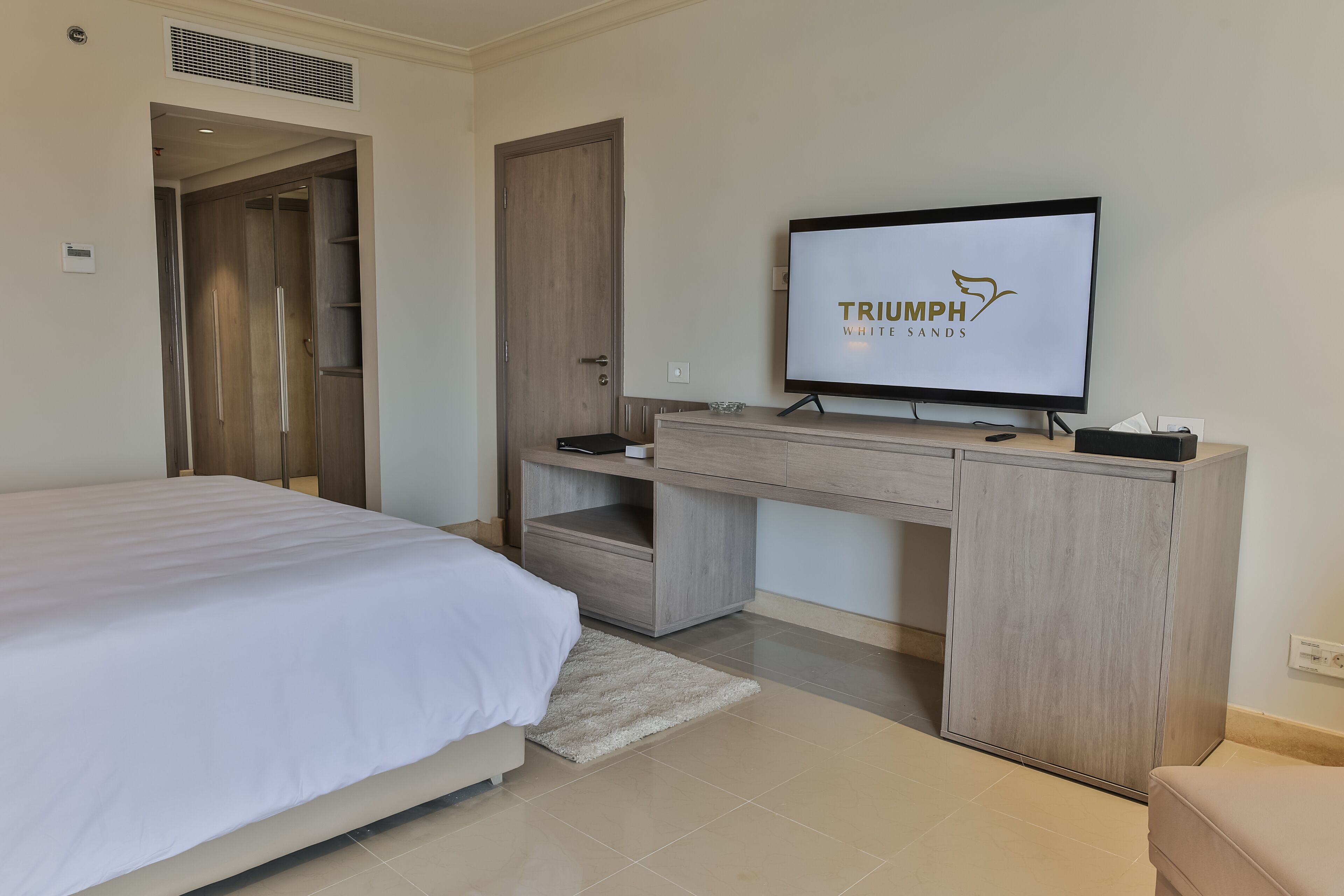 Ein modernes Hotelzimmer mit einem Bett, einem Schreibtisch mit Computer und einem Fernseher, der 'TRIUMPH WHITE SANDS' zeigt.
