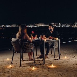 A couple dining at a table with candles on a beach at night, overlooking a city skyline.