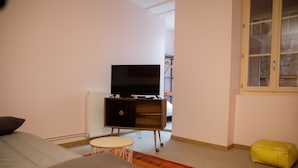 Un dormitorio con una cama, un televisor sobre un mueble de madera, una mesita redonda y un reposapiés amarillo.