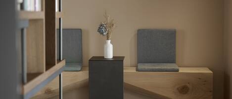 A modern seating area with a dark square table, a white vase with dried flowers, and two grey upholstered chairs against a plain wall.