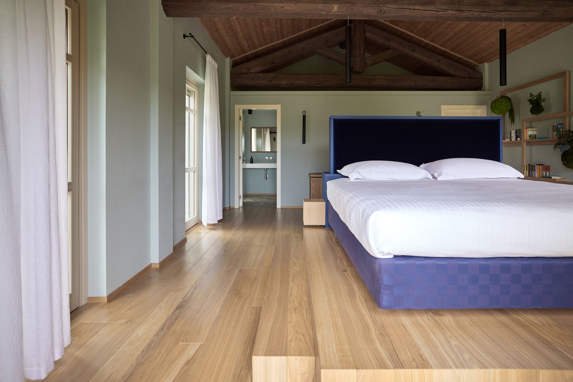 A bedroom with a large bed, wooden flooring, a high ceiling with exposed beams, a small bedside table, and a nightstand.